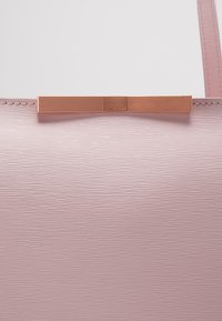 Gros plan d’un sac à main texturé rose pâle avec une fine bandoulière et un détail barre métallique horizontale en or rose en haut au centre.