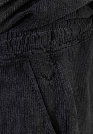 Közeli kép fekete bordázott szövetnadrágról, begyűrt rugalmas derékpánttal és kis hímzett logóval az oldalzseb közelében.