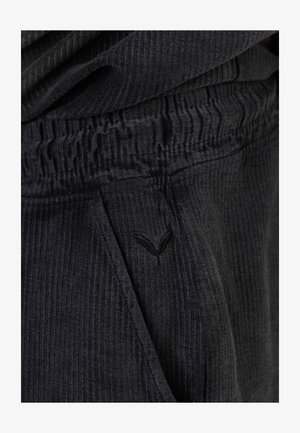 Közeli kép fekete bordázott szövetnadrágról, begyűrt rugalmas derékpánttal és kis hímzett logóval az oldalzseb közelében.