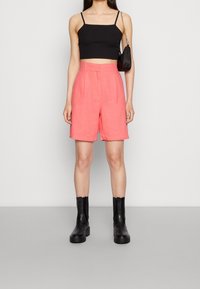 Černý zkrácený top, růžové šortky s vysokým pasem a předními záhyby a černé kotníkové boty s silnými podrážkami. Jednoduché pozadí.