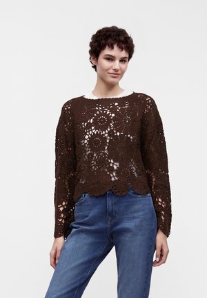 Ung person med kort krøllet hår iført en brun hæklet sweater over en hvid top og blå jeans, stående mod en ensfarvet lys baggrund.
