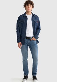 Camisa de mezclilla en azul oscuro con dos bolsillos en el pecho, parte delantera abotonada y detalles de costura. Combinada con jeans azul claro y zapatillas negras.