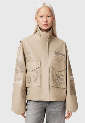 Nainen, jolla on suoraksi käsitellyt vaaleat hiukset, beige nahkatakki, jossa korkea kaulus ja useita kirjailtuja kuvioita, yhdistetty vaaleanharmaisiin farkkuihin.