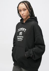 Čierna mikina s kapucňou s predným vreckom, veľkým bielym nápisom "TOMMY 85 AUTHENTIC ORIGINAL" a rebrovanými manžetami. Hladká bavlnená tkanina, voľný strih.