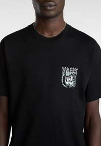 Sort bomuld t-shirt med et grafisk tryk på venstre bryst, der viser et skelet omgivet af vinstokke i hvid og lys grøn.