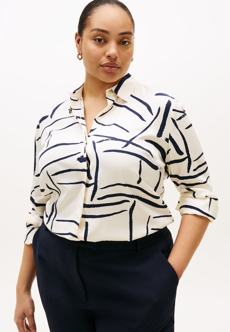 Λευκό πουκάμισο με κουμπιά μπροστά, με σκούρο μπλε αφηρημένο σχέδιο πινελιές, μανίκια που μπορούν να γυριστούν επάνω και ελαφρύ ύφασμα. Συνδυάζεται με σκούρο μπλε παντελόνι.