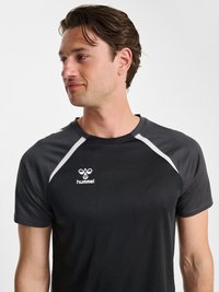 Junger Mann mit braunen Haaren, der ein schwarzes Sport-T-Shirt mit weißem Rand und weißem Hummel-Logo trägt und nach links schaut vor weißem Hintergrund.
