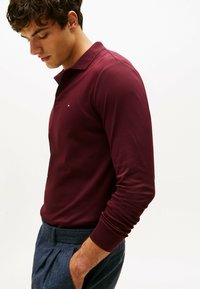 Burgunderfarbener Langarm-Polo-Shirt mit Kragen, ausgestattet mit einem kleinen Logo auf der linken Brust. Das Modell trägt maßgeschneiderte graue Hosen.