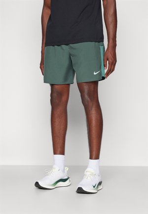 Mężczyzna w zielonych sportowych szortach Nike, czarnej koszulce, białych skarpetach i białych butach do biegania Nike na jednolitym tle.