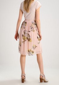 Jupe florale rose clair avec ceinture froncée, ornée de motifs de fleurs jaunes et grises. Portée avec un haut ajusté clair et des sandales à talons.