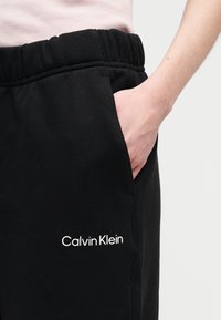 Χέρι ατόμου στην τσέπη μαύρου παντελονιού φόρμας Calvin Klein με ελαστική μέση, που φοράει ανοιχτόροζ μπλουζάκι, σε ουδέτερο φόντο.