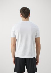 Bílý krátký rukáv tričko vyrobené z bavlny, s kulatým výstřihem a hladkou texturou, nošeno s černými šortkami, pohled zezadu.