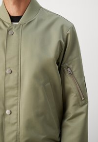 Homme portant une veste bomber vert clair avec boutons-pression et poche zippée sur la manche gauche supérieure sur fond uni.