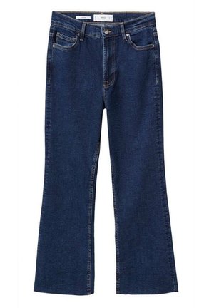 Vrouw die donkerblauwe high-waist jeans, een grijze off-shoulder cropped trui en zilveren puntige hakken draagt, staand op een simpele vloer.