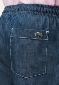 Κοντινό πλάνο σε μπλε τζιν παντελόνι με ελαστική μέση, πίσω τσέπη και πράσινο λογότυπο κροκόδειλου στη γωνία της τσέπης.