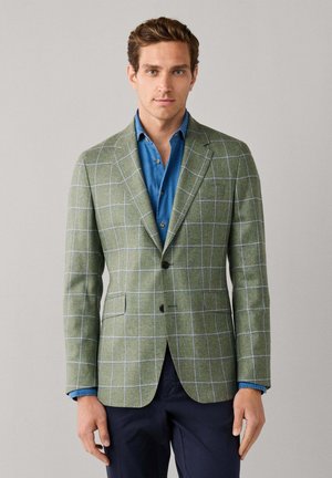 Hombre con blazer verde de cuadros ventana sobre camisa azul y pantalones azul marino, de pie frente a un fondo claro y liso.