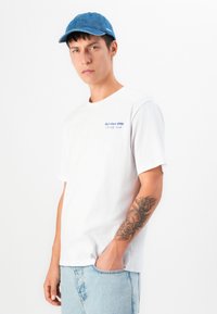 White cotton t-shirt with blue text "J&J since 1990" on chest, paired with a blue denim cap. Model stands with hand in pocket, showing arm tattoo.