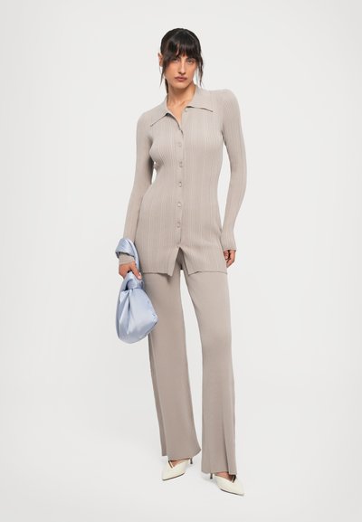 Woman wearing beige ribbed button-up cardigan and matching pants, holding a light blue satin handbag, standing against a white background.