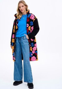 Čierny kabát s kvetinovým motívom a texturovanou tkaninou, skombinovaný s modrým svetrom a širokými džínsami. Čierne platformové topánky.
