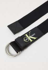 Black elastic belt with a leather tag featuring "calvinKlein" in green, silver metal D-ring closure, and textured accents.