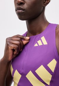 Άτομο με σκούρο δέρμα που φοράει μωβ αθλητική μπλούζα αποσυμπλέκοντας το γιακά, αποκαλύπτοντας μπεζ γεωμετρικά σχήματα στο ύφασμα.