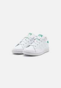 White sneakers made of synthetic leather, featuring green accents, perforations on the side, and a rubber outsole. Laces and a cushioned collar.