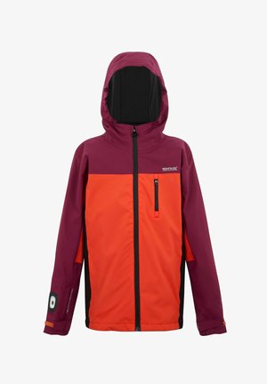 Młody chłopiec uśmiechający się, ubrany w kurtkę w blokach kolorów pomarańczowo-bordową, czarne spodnie, czarne buty i wzorzysty komin na szyję, stojący na jednolitym tle.