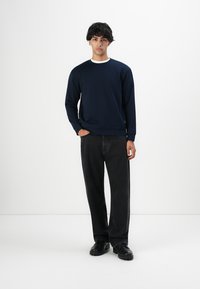 Joven de pie vestido con una sudadera navy, unos vaqueros negros de pierna ancha y zapatos negros frente a un fondo blanco.