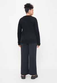 Bluză neagră cu mâneci lungi și croială lejeră, cu pantaloni negri cu picioare largi și pantofi negri masivi. Design simplu, fără modele vizibile.