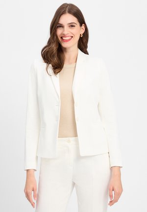 Blazer blanc sur mesure avec une fermeture à deux boutons, des revers en pointe et des poches avant, associé à un haut beige et un pantalon blanc assorti.