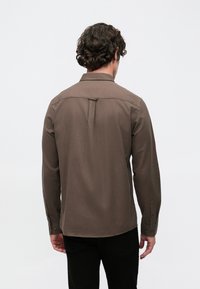 Braunes Langarmhemd mit klassischem Kragen und geknöpften Manschetten. Der Rücken zeigt eine ausgeprägte Naht und eine Kellerfalte als zusätzliches Designelement.