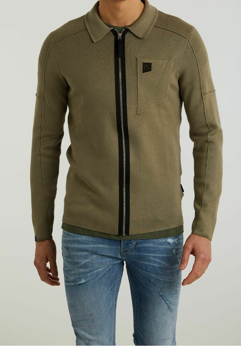 Homme portant un pull zippé vert olive avec col et petite poche poitrine, associé à un jean bleu clair déchiré.