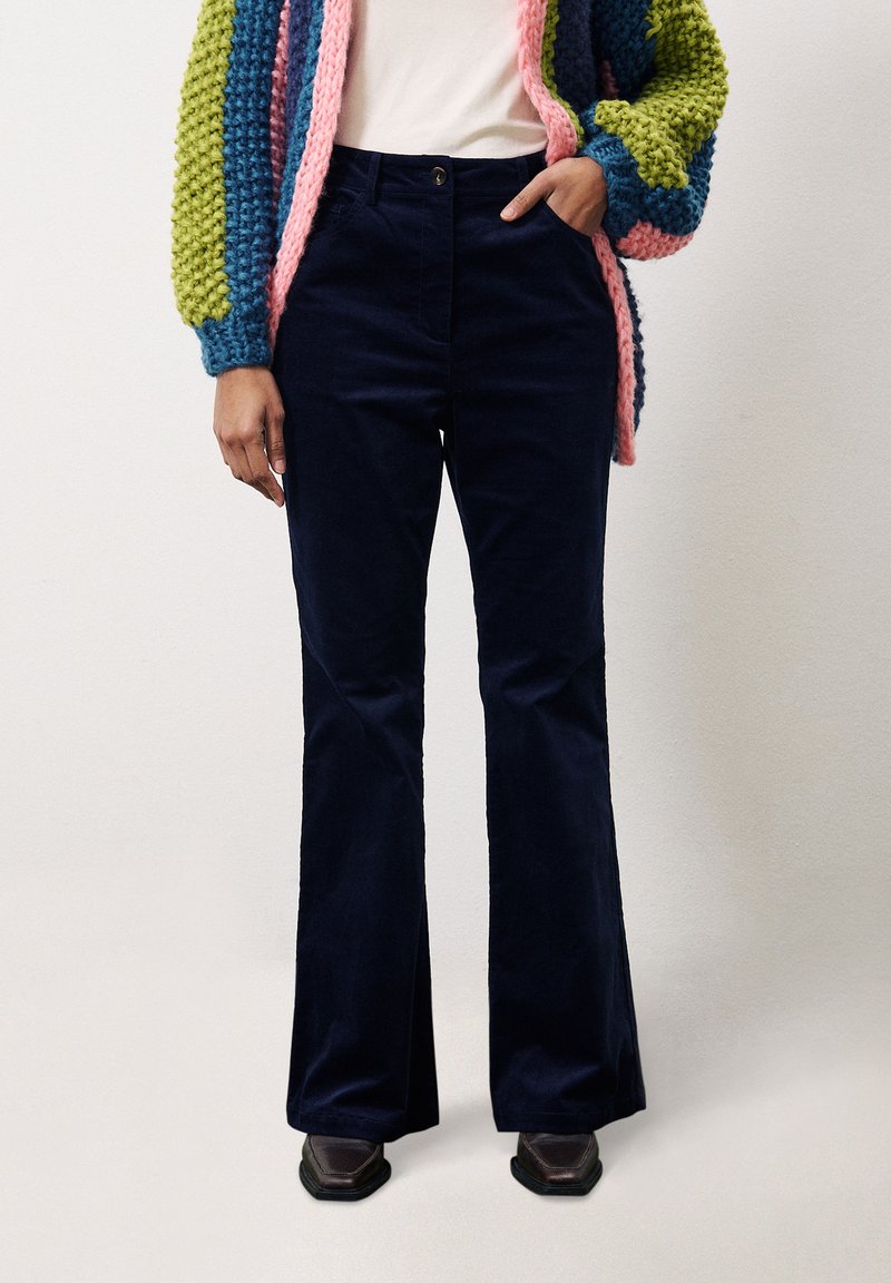 Marineblaue Cordhosen mit weit auslaufendem Bein und hoher Taille, verstärkten Nähten und einer vorderen Knopfschließe. Getragen mit einer bunten Strickjacke.