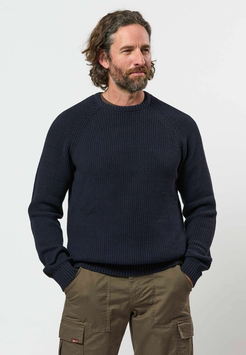 Homme aux cheveux bouclés et à la barbe, portant un pull en laine bleu foncé et un pantalon cargo marron, debout les mains dans les poches sur un fond uni.