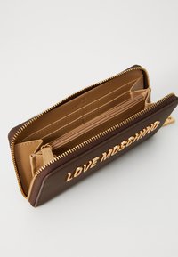 Rudas odinis piniginė su auksiniais aksesuarais. Jame yra užtrauktukas, keli kortelių laikikliai ir logotipas išspaudintas priekyje. Vidus yra smėlio spalvos.
