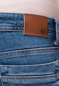 Πίσω τσέπη από μπλε τζιν με διπλή ορατή ραφή και καφέ δερμάτινο ταμπελάκι στη μέση, πάνω σε ανοιχτό ροζ ύφασμα.