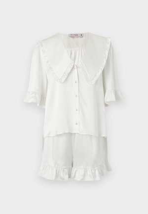 Λευκή μπλούζα με κοντά μανίκια, κουμπιά και μεγάλο βολάν γιακά με ταιριαστά σορτς με βολάν στα τελειώματα, εμφανίζεται σε απλό φόντο.