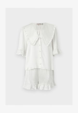 Λευκή μπλούζα με κοντά μανίκια, κουμπιά και μεγάλο βολάν γιακά με ταιριαστά σορτς με βολάν στα τελειώματα, εμφανίζεται σε απλό φόντο.