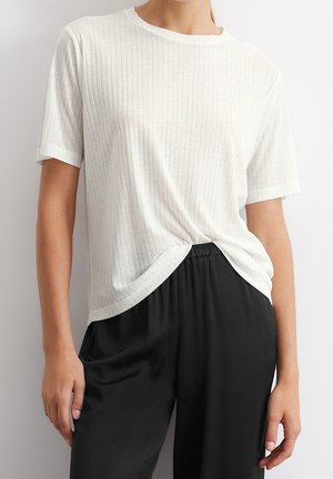 Persoon draagt een wit geribd T-shirt met korte mouwen, gestopt in losse zwarte broek, staand tegen een effen witte achtergrond.