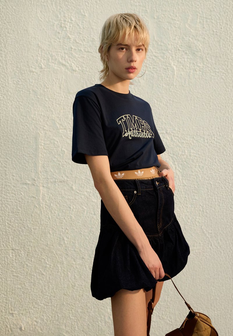 Navy t-shirt with "TIMBS" graphic, paired with a dark denim skirt featuring a flared hem, and a brown bag with quilted texture.