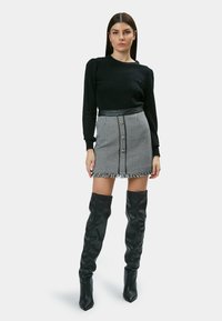 Schwarzer strukturierter Pullover mit langen Ärmeln, kombiniert mit einem Minirock im Hahnentrittmuster mit Fransenabschluss und vorderen Knöpfen. Schwarze Overknee-Stiefel.