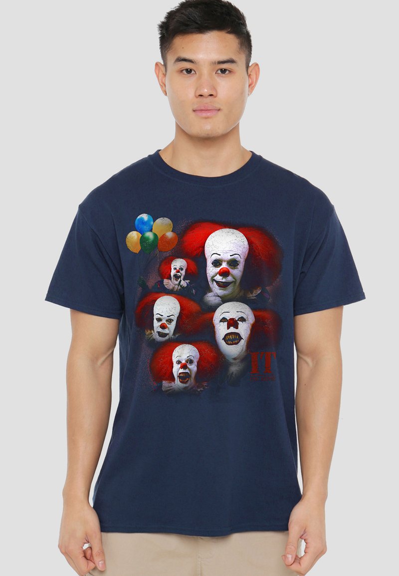 Young man wearing navy shirt with graphic of five red-haired clown faces, colorful balloons, and "IT" text in red on front.