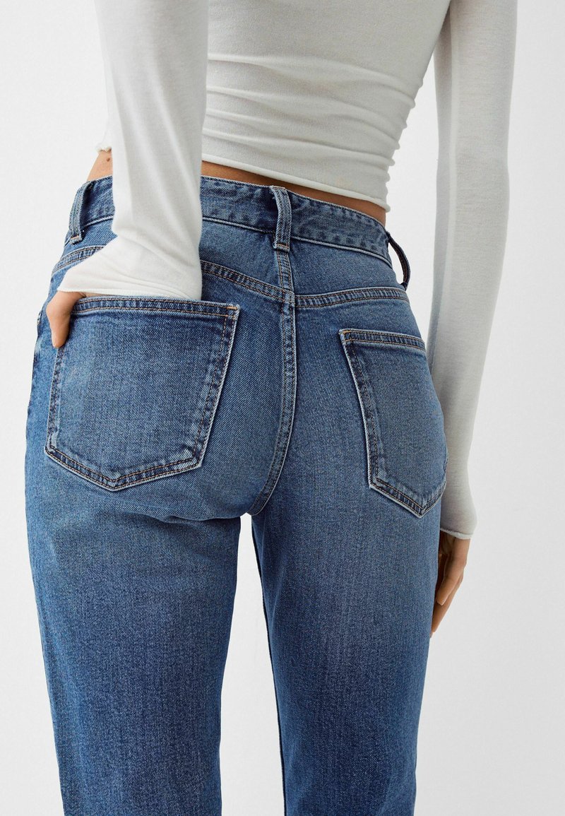 Jeans in denim blu con vita alta, taglio dritto e due tasche posteriori. Il tessuto presenta una texture classica con dettagli di cucitura visibili.