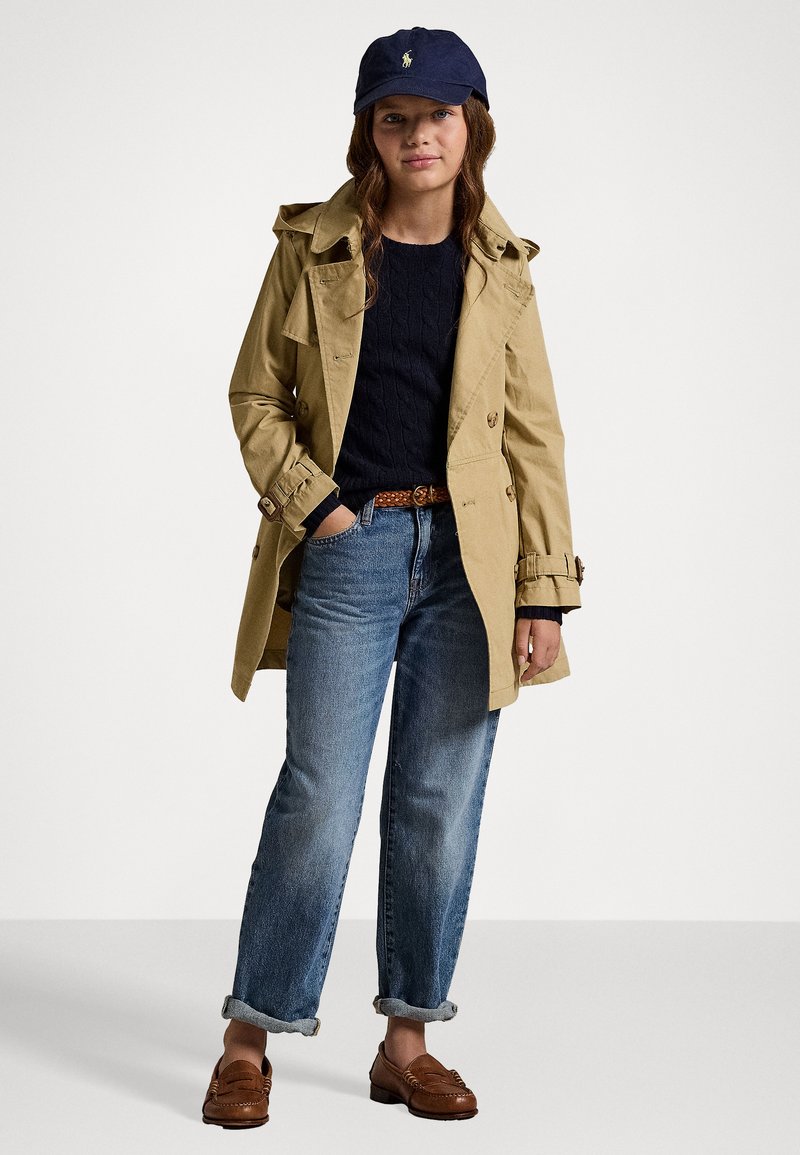Beige trenchcoat med dobbelspent knapper, marineblå strikkegenser, rullende lyseblå jeans, brune loafers, og en marineblå caps.