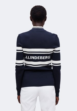 Personne portant un pull bleu marine avec des rayures blanches et le texte "J.LINDEBERG" dans le dos, associée à un pantalon blanc, dos au spectateur.