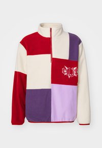 Fleece pullover med halvlynlås, prydet med et patchwork-design i rød, hvid, lilla og lys lilla, fremhævet af et logo på brystet.