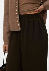 Cardigan marron en tricot avec des boutons semblables à des perles, haut beige et pantalon plissé marron foncé avec une taille élastique. La texture semble douce.