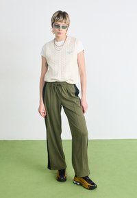 Krämfärgad kabelstickad ärmlös tröja över en vit t-shirt, kombinerad med lösa olivgröna träningbyxor med svarta sidosträck och en logo. Svarta och gyllene sneakers.
