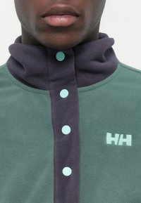 Fleece pullover in teal met een donkerpaarse kraag, met lichtblauwe knopen en een wit logo op de linkerborst. Zachte textuur.