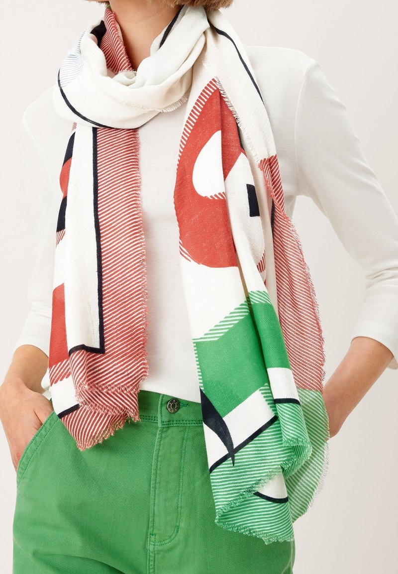 Personne portant une grande écharpe blanche avec des motifs abstraits rouges, verts et noirs sur un haut à manches longues blanc et un pantalon vert.